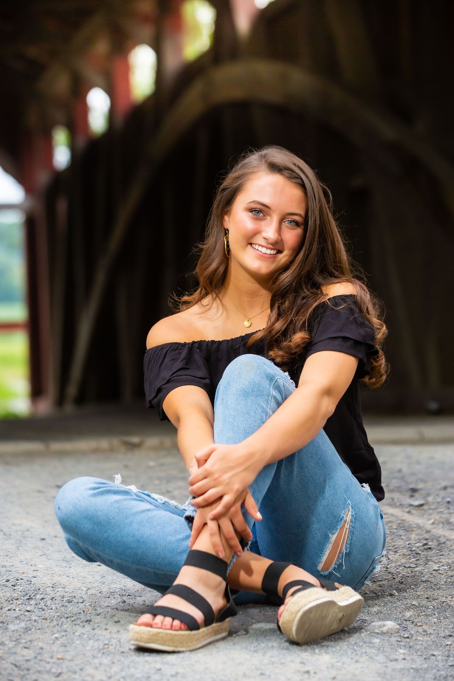 Casual portrait sitting cross-legged wearing off-shoulder top and distressed jeans under covered bridge.