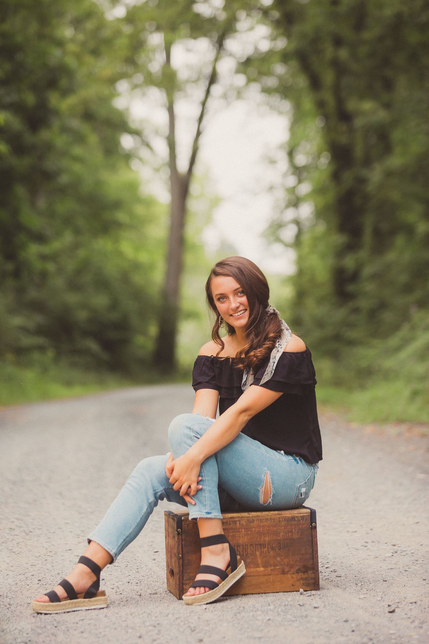 A casual portrait on a dirt path with someone sitting on a wooden crate wearing distressed jeans and sandals.