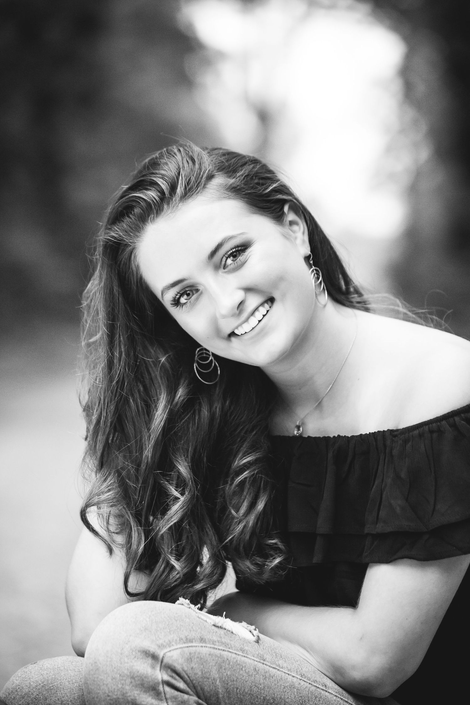 Black and white portrait with off-shoulder top and long wavy hair against blurred nature background.