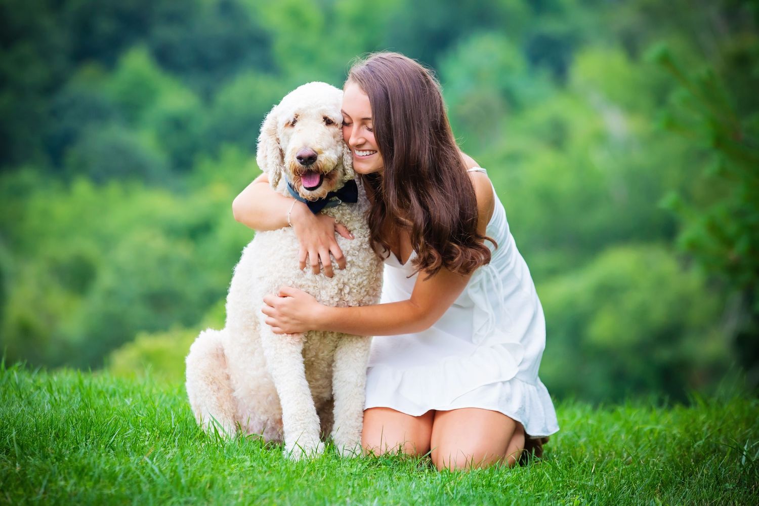 Person in white dress hugging and playing with a friendly golden doodle dog on green grass.