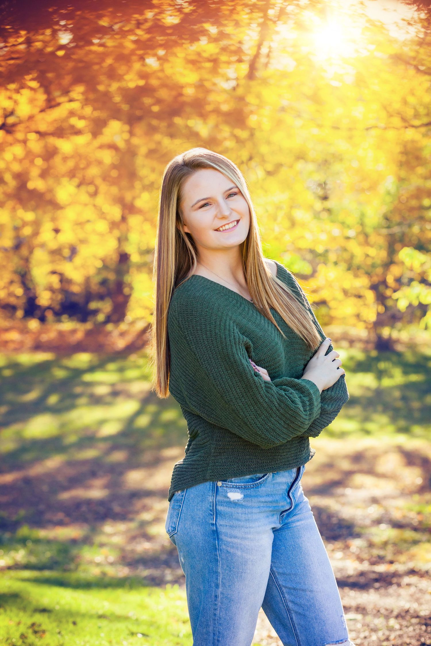 Outdoor portrait in autumn foliage wearing green sweater and jeans with golden sunlight.