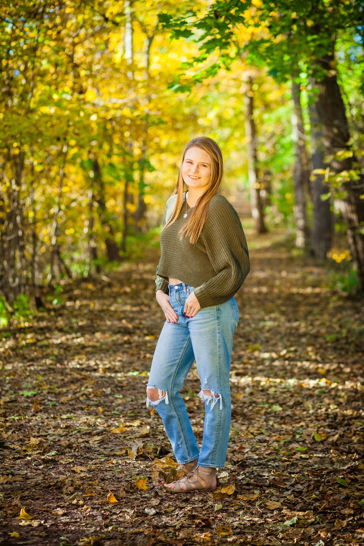 Fall portrait in a forest with someone wearing ripped jeans and olive sweater among yellow leaves.