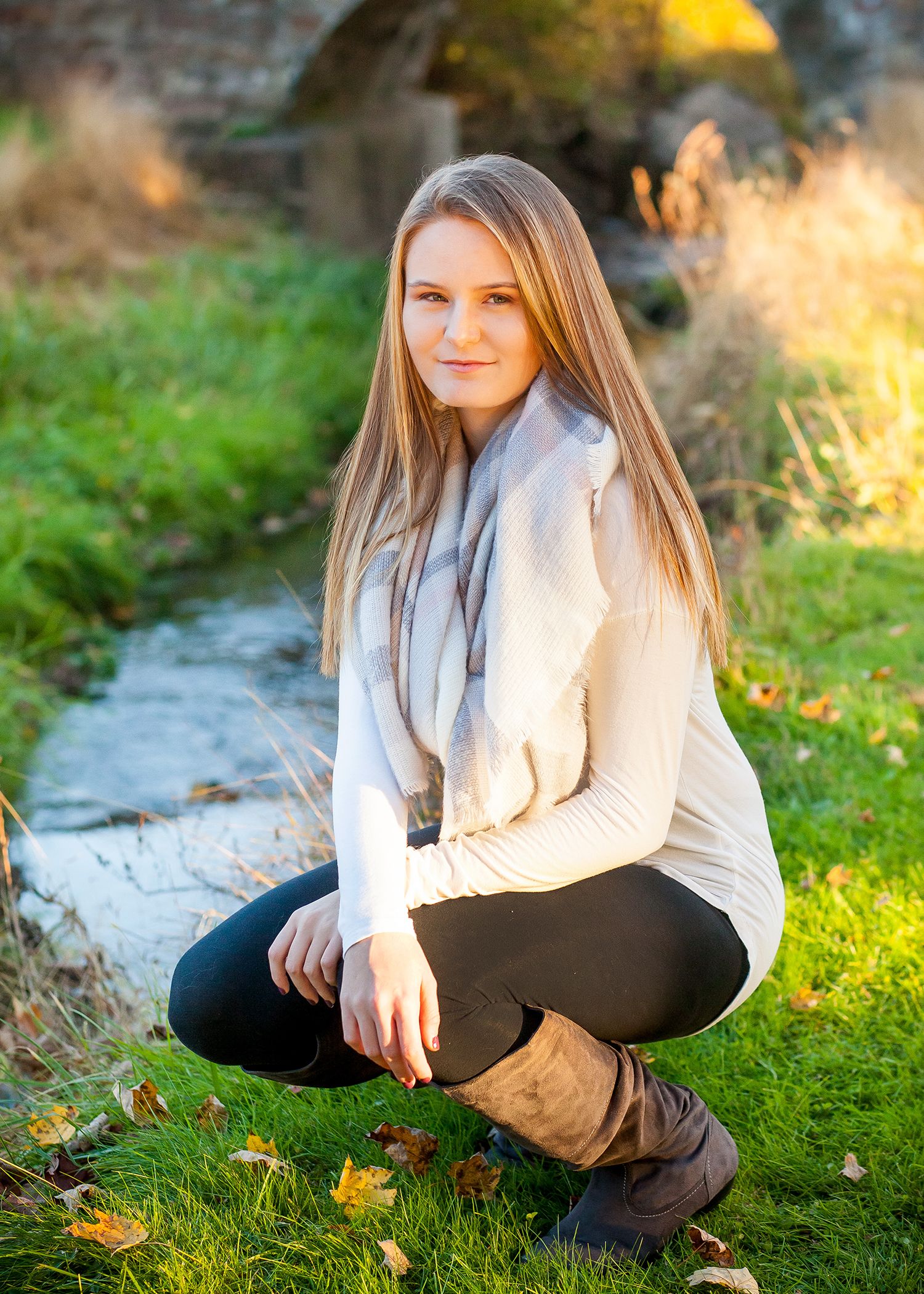 Autumn portrait by a creek wearing cream sweater and boots while crouching on grass.