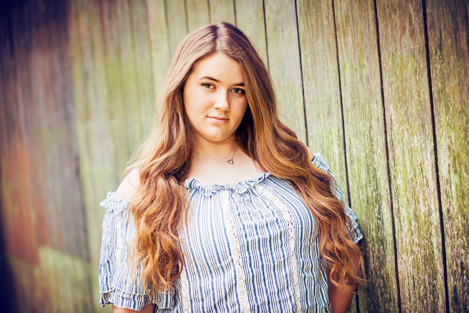 Portrait against wooden fence wearing striped peasant top with long wavy hair.