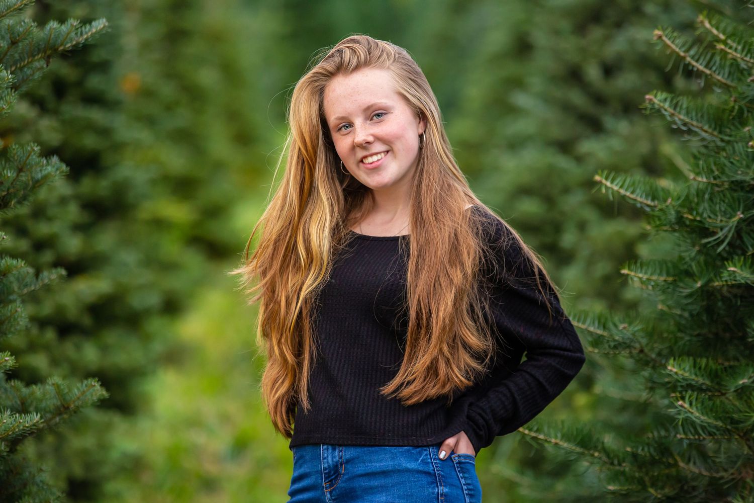 Portrait of someone in a black sweater and jeans standing outdoors near evergreen trees.