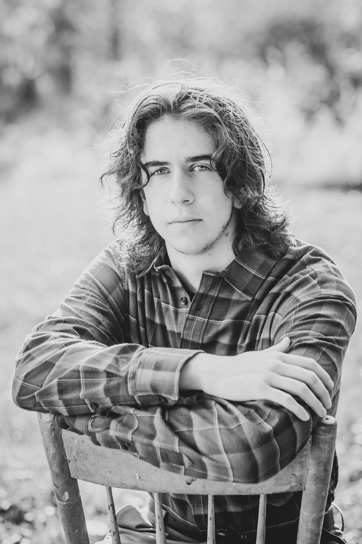 Black and white portrait of someone with long hair wearing plaid shirt while sitting backwards on chair.