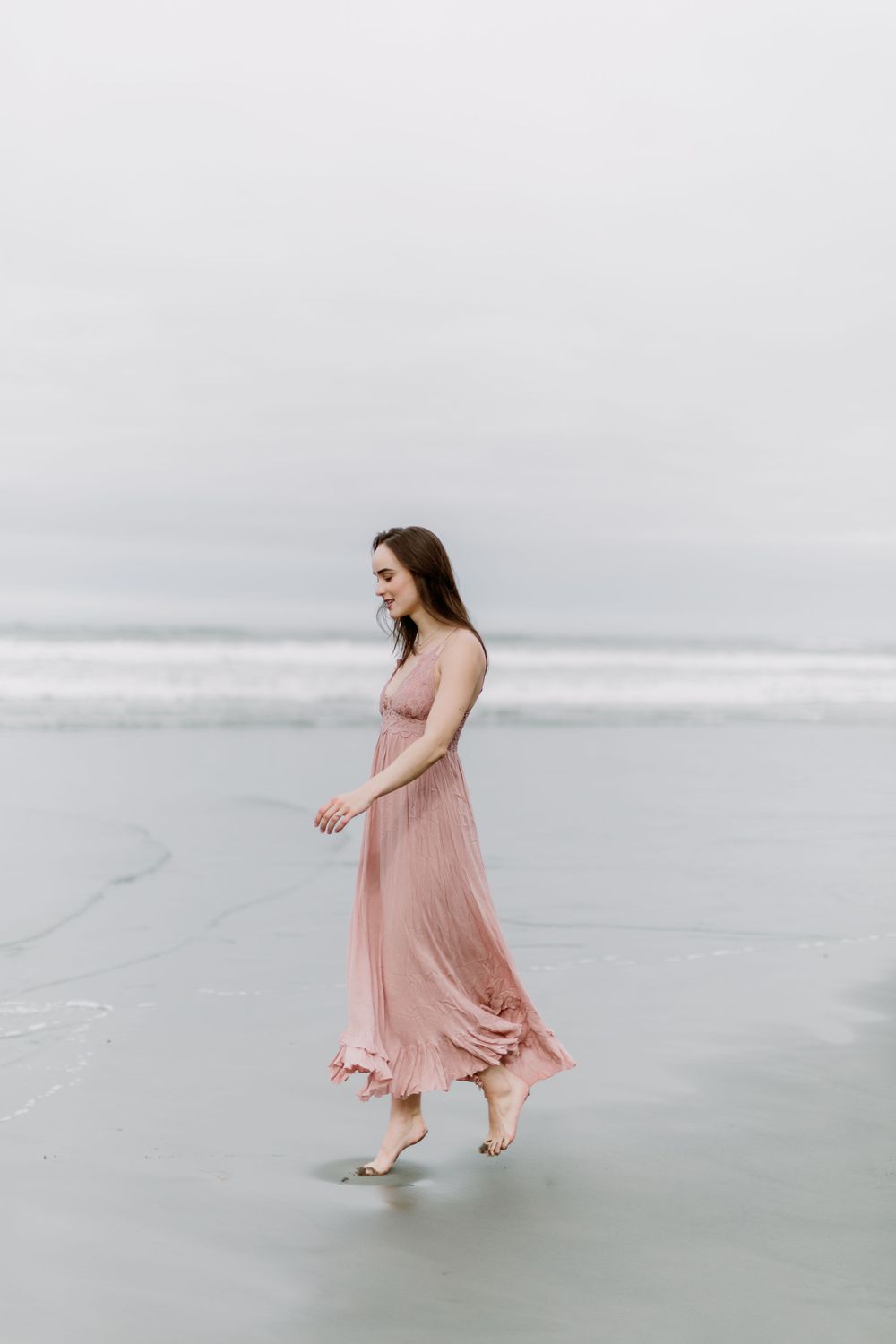 Woman in flowing pink dress walking alone on misty beach with waves in background.