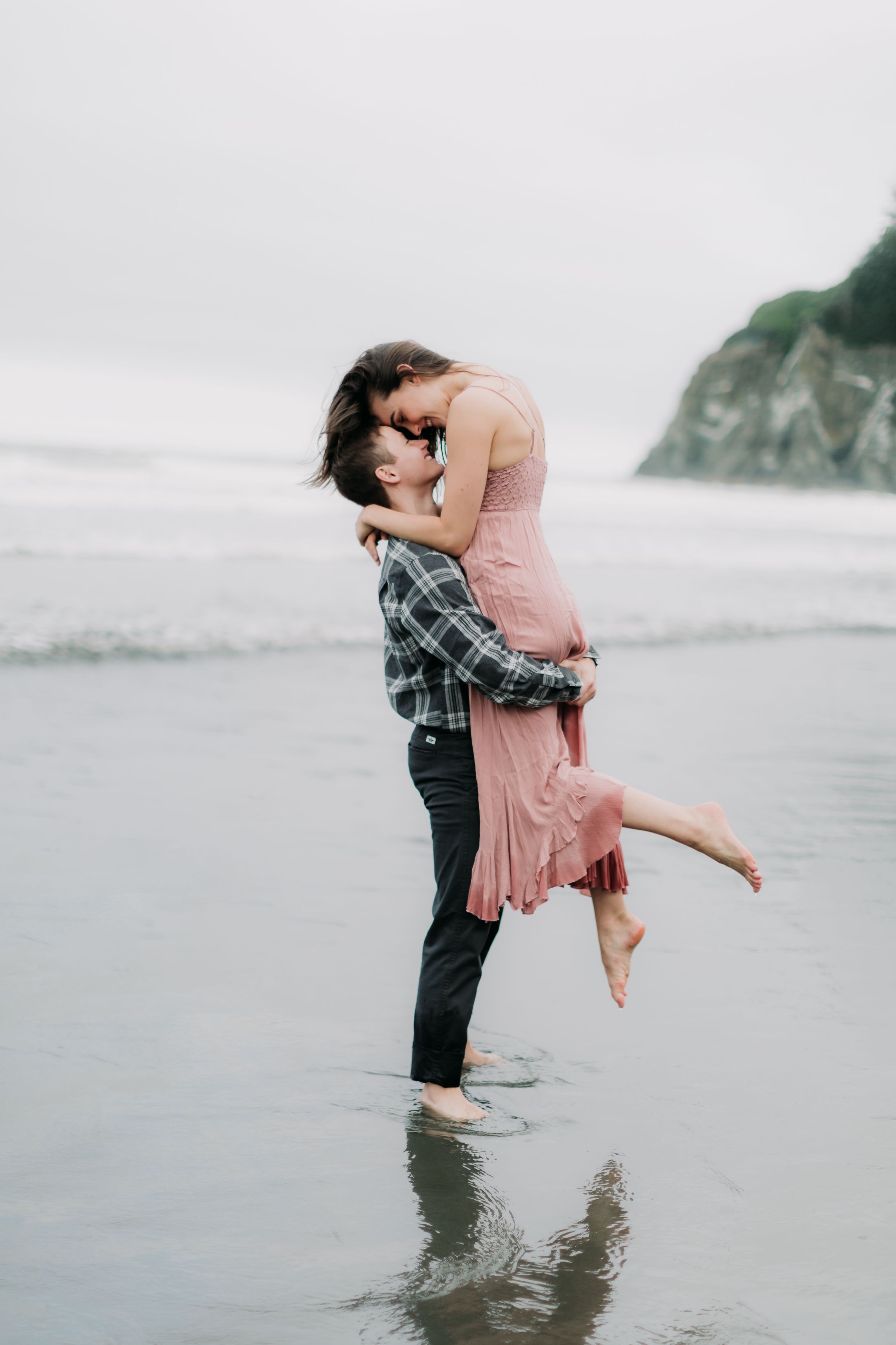 Romantic beach scene with couple embracing in shallow water against misty coastal cliffs.