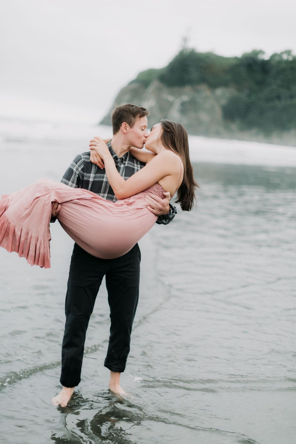 Couple sharing romantic moment in shallow ocean water with coastal cliffs in background.