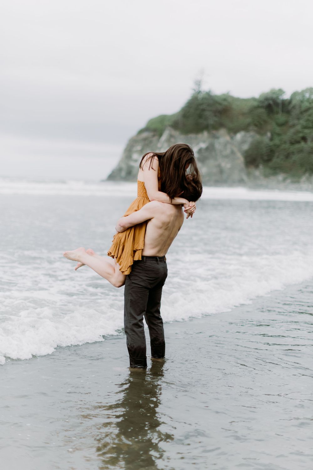 Romantic beach embrace with waves and coastal cliffs in background.