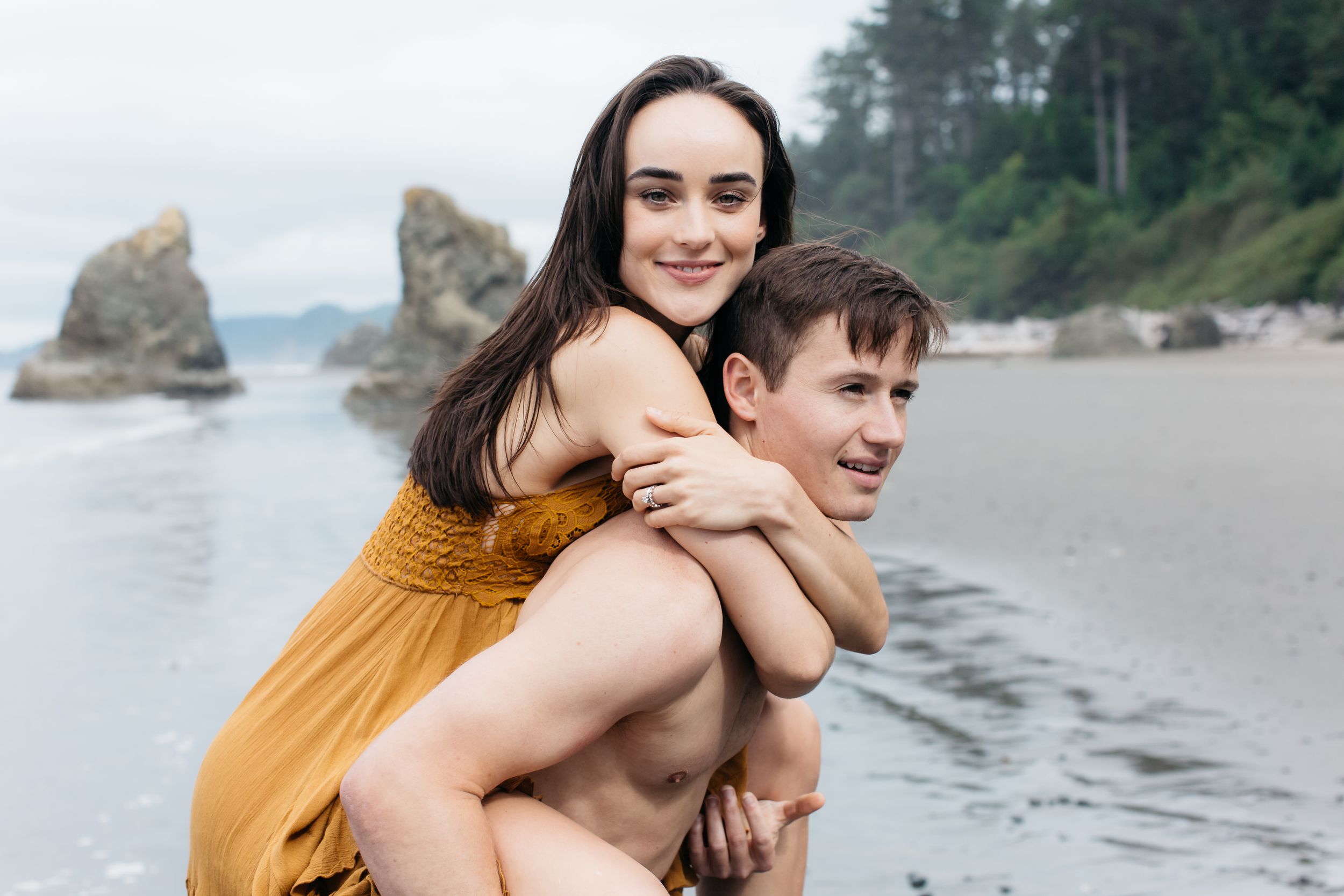 A playful piggyback ride on a scenic beach with dramatic coastal rock formations in the background.