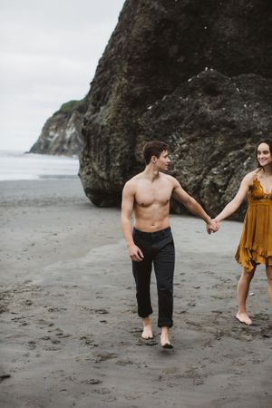 A couple walks hand in hand on a dramatic black sand beach with towering cliffs.