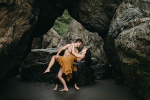 A romantic couple pose together under a natural rock arch on a dark sandy beach.