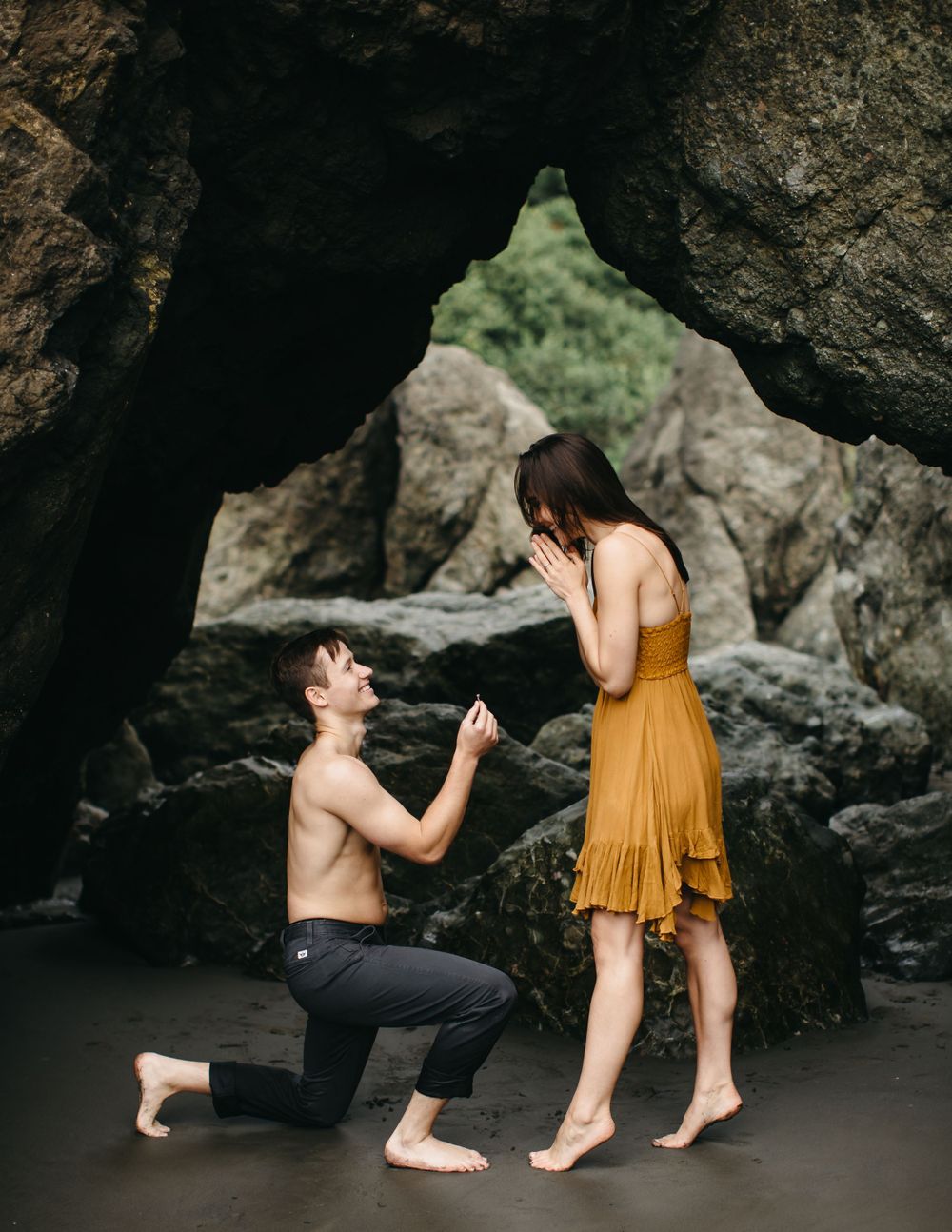A surprise proposal moment captured under a dramatic rock formation on the beach.