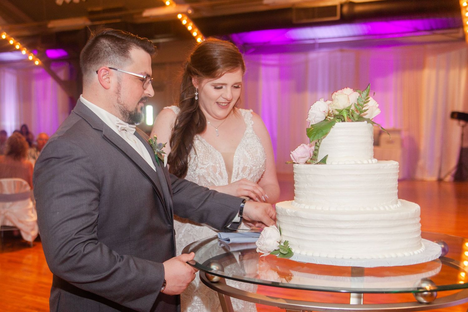 Wedding couple cutting their elegant white tiered cake under purple and orange venue lighting.
