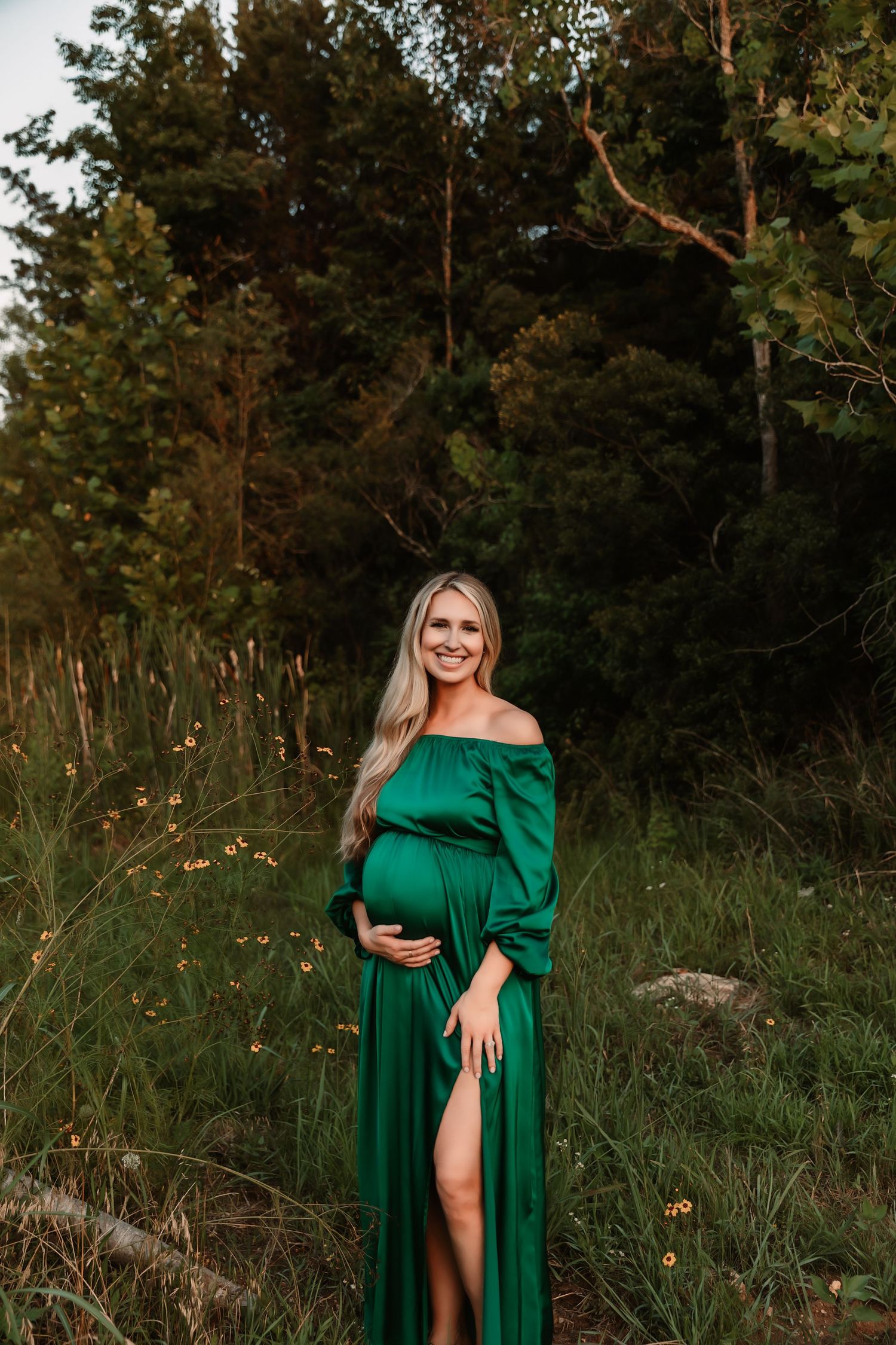 Maternity photo of woman in elegant green off-shoulder dress against forest backdrop.