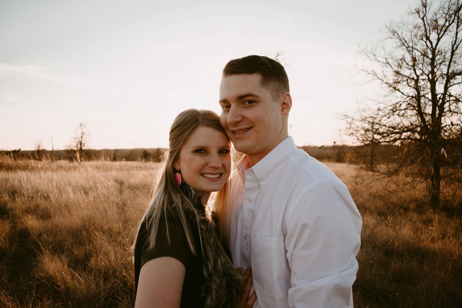 A romantic couple embraces in a field at sunset with golden light and bare trees in the background.