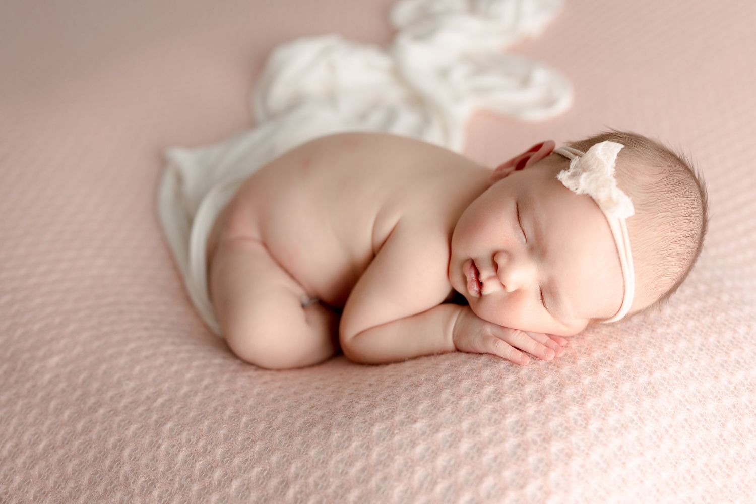A sleeping newborn baby in white lace wrap poses on pink textured backdrop.