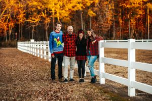 A family poses together by a white fence during autumn with colorful fall foliage behind them.