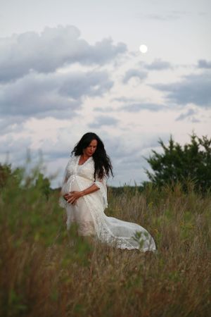 A maternity photo of a woman in white lace dress standing in tall grass at dusk.