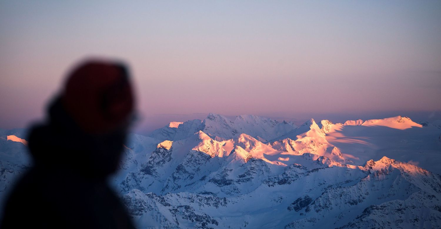 Silhouette of a person gazing at snow-capped mountain peaks glowing pink at twilight.