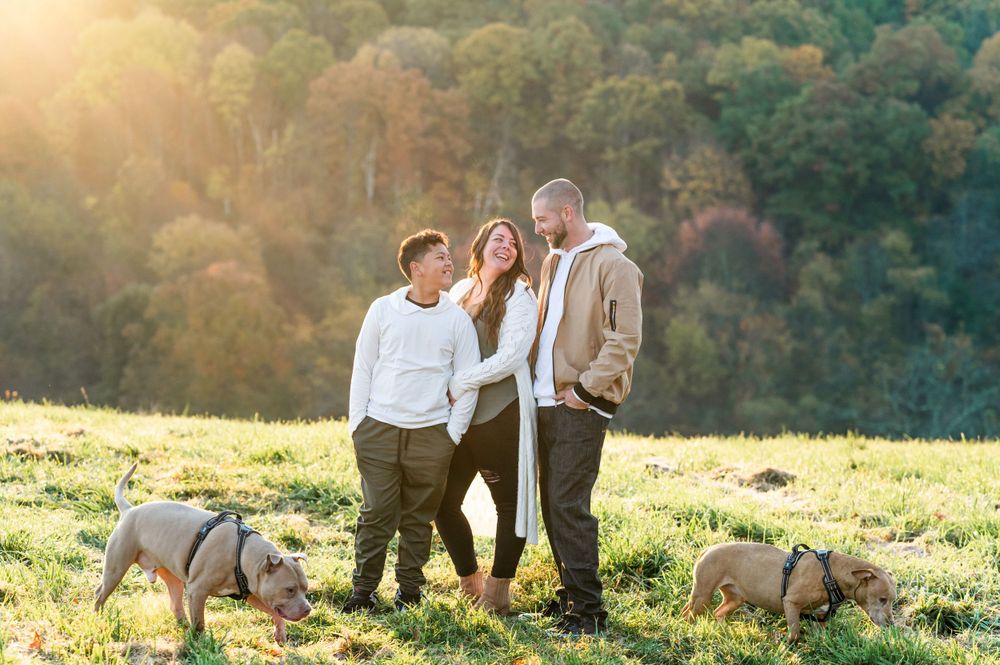 Family and two dogs pose together on sunny hillside with autumn trees in background.