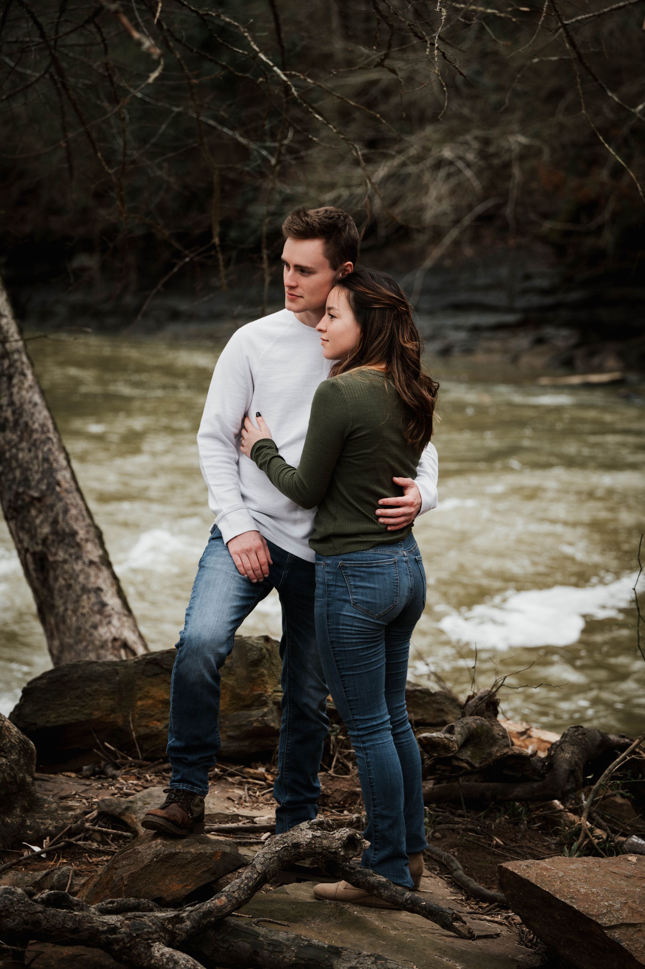Couple embracing by river's edge surrounded by winter trees and rocks.