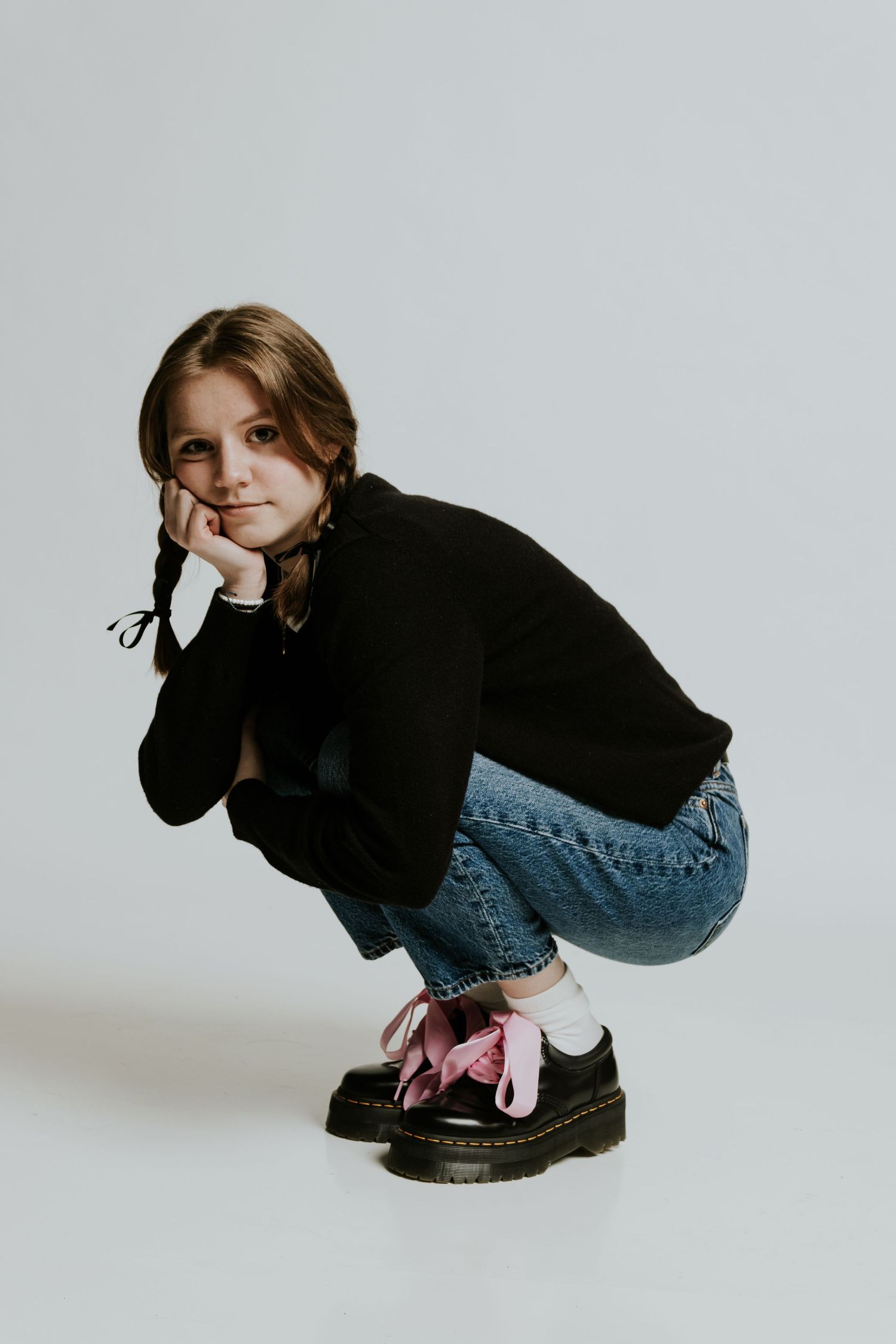 Artistic portrait of someone in black sweater and jeans crouching against white background.