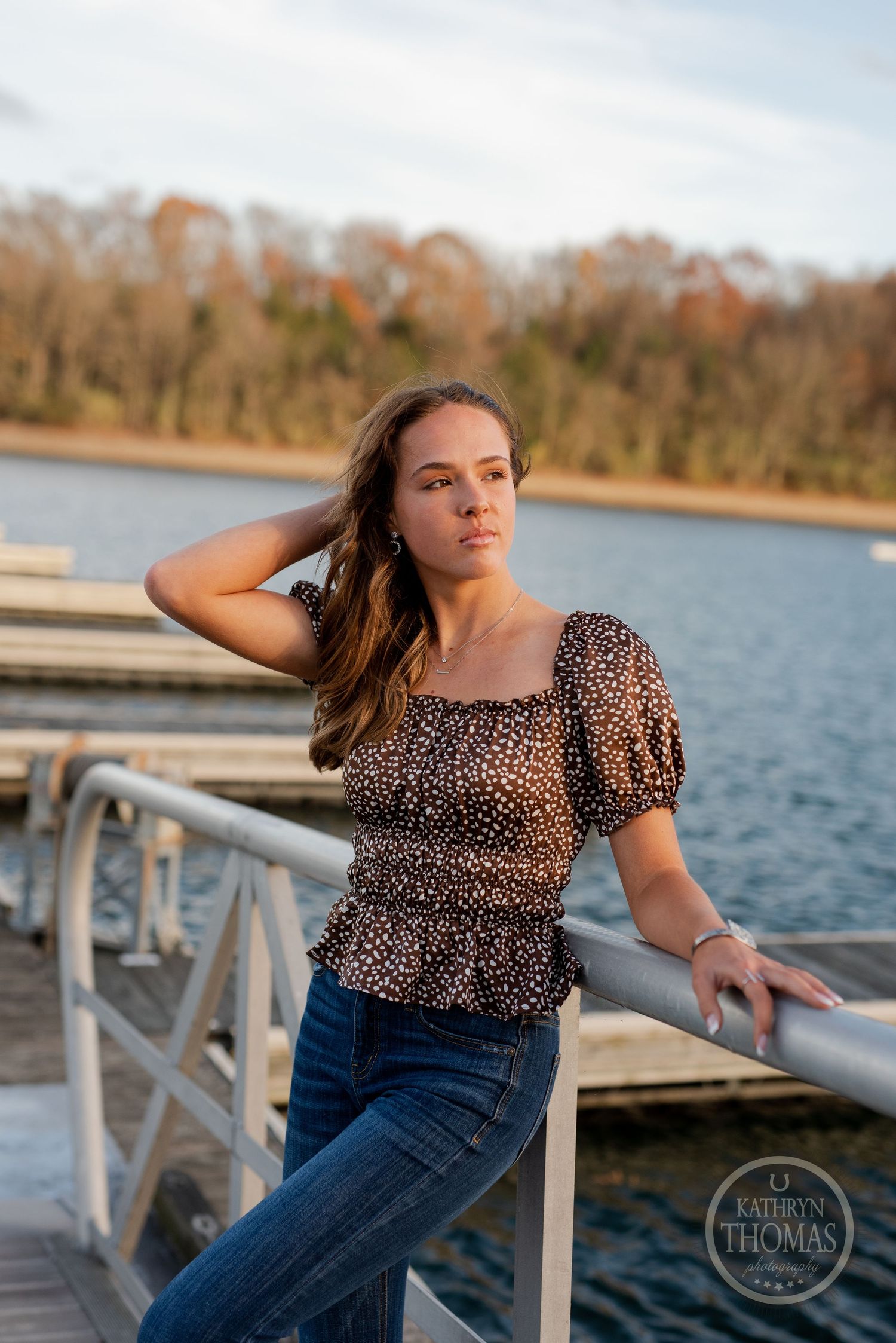 Portrait of a person in a floral puff sleeve top leaning on a dock railing at sunset with autumn trees in background.