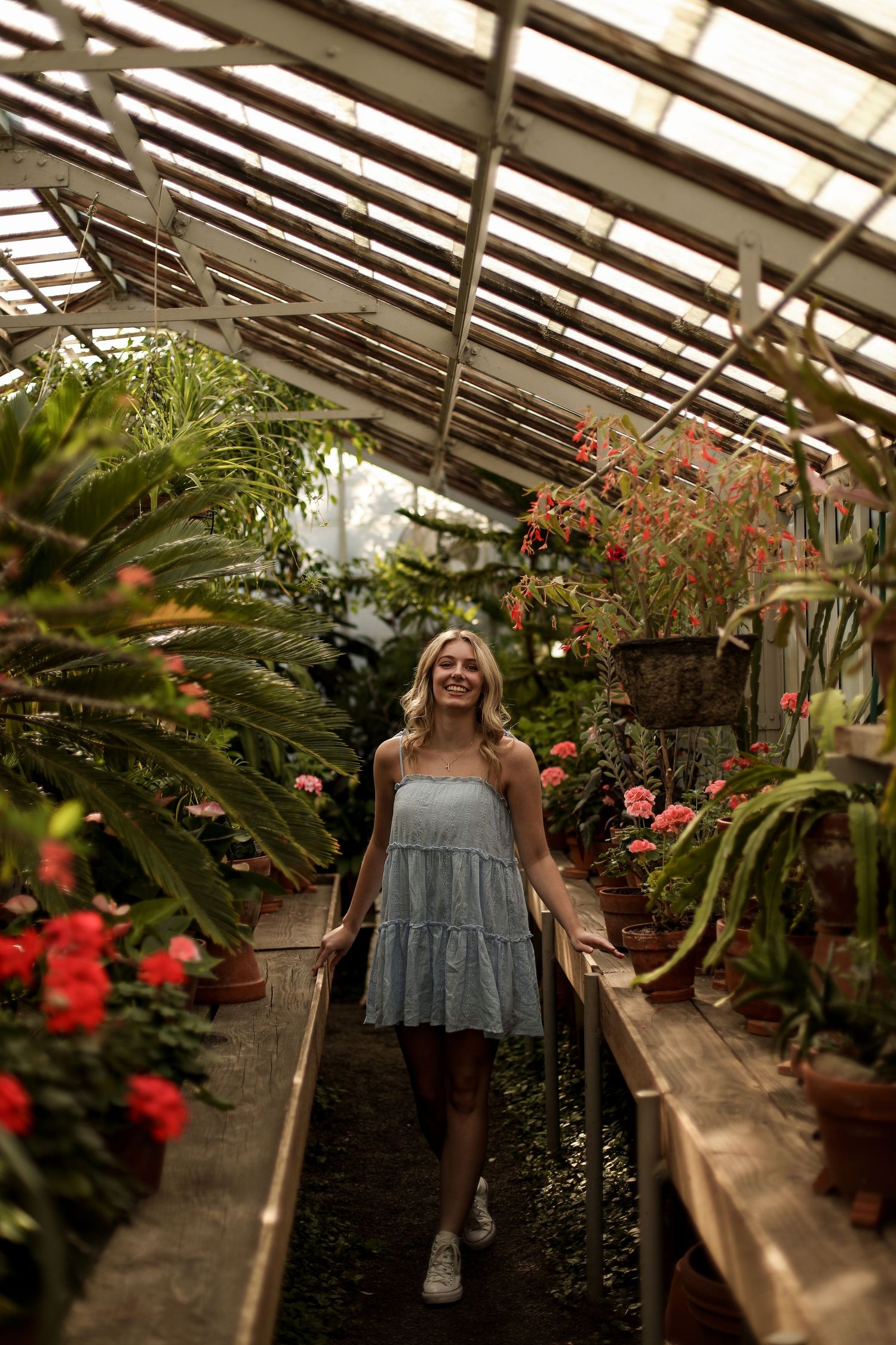 Young person in a flowy dress walks through a greenhouse filled with tropical plants and colorful flowers.