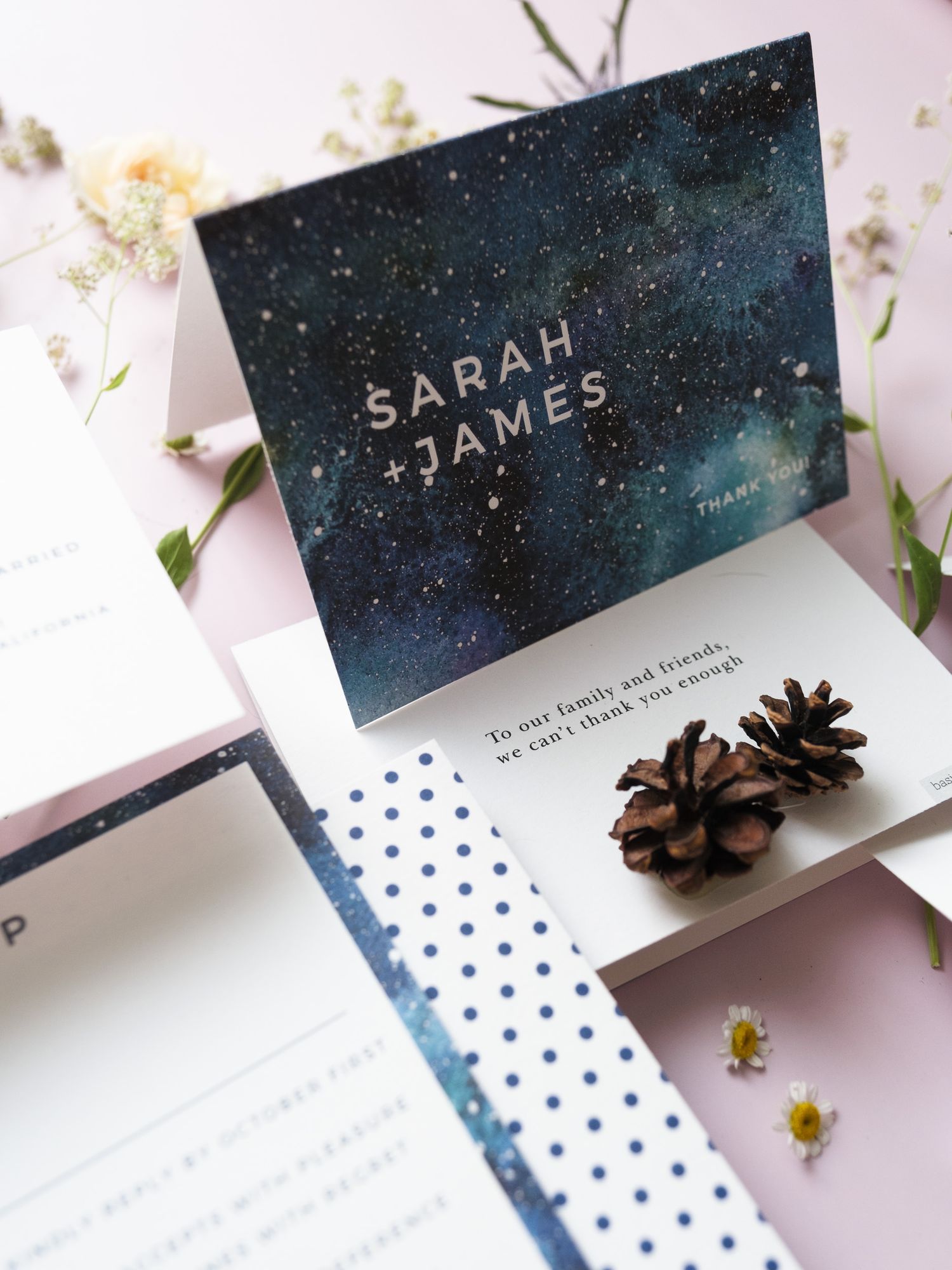 Wedding invitation with galaxy design and names Sarah + James on navy blue card with matching polka dot details and pinecones.