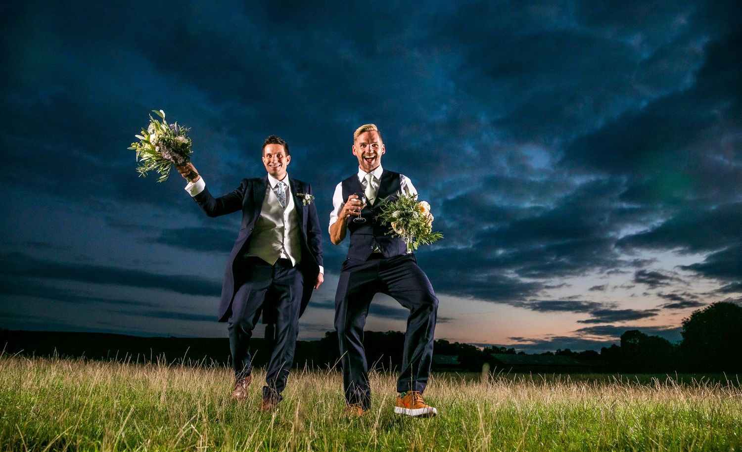 Two groomsmen jump playfully in a field at sunset holding bouquets against dramatic sky.
