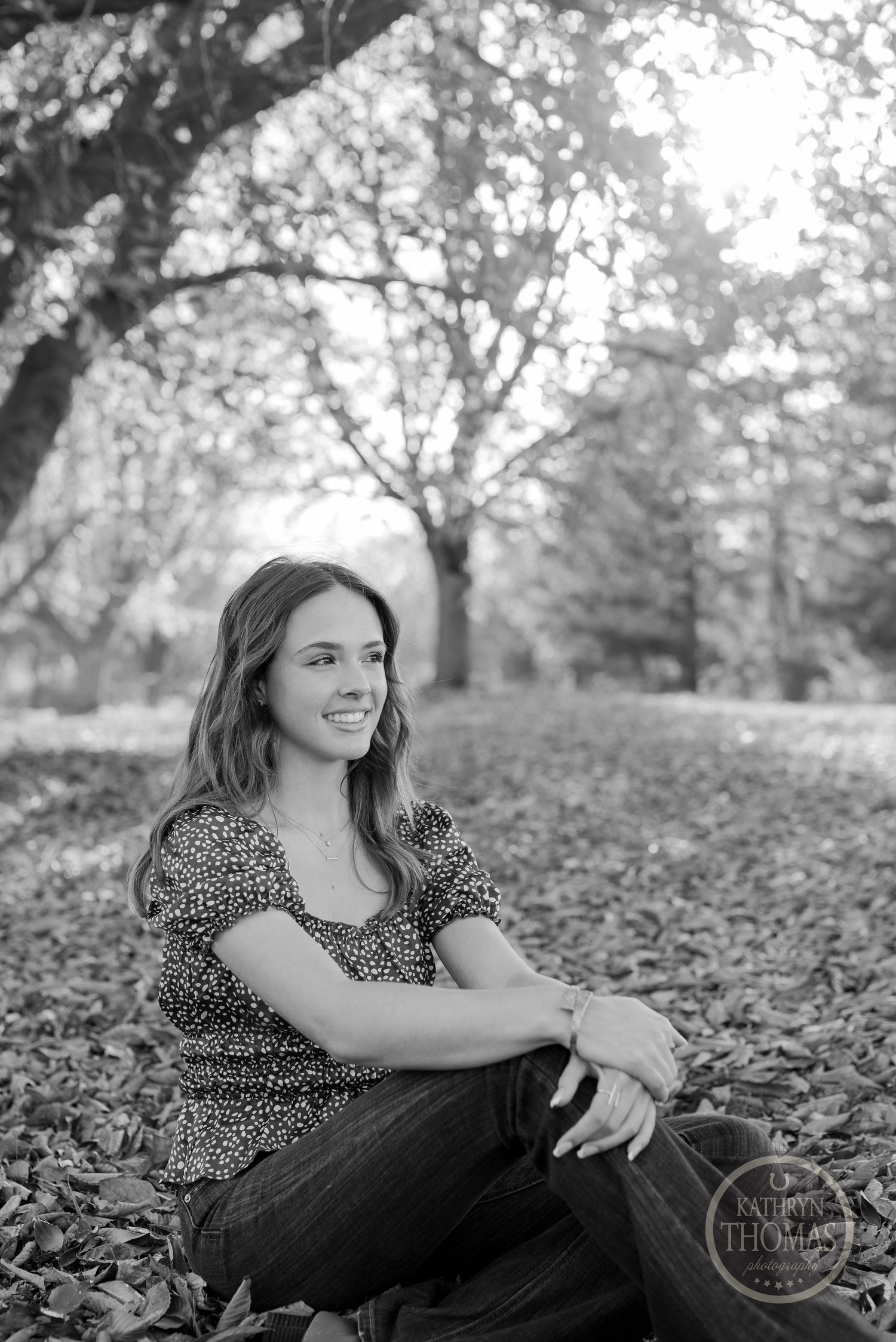 A black and white portrait sitting on fallen leaves wearing a patterned top and jeans.