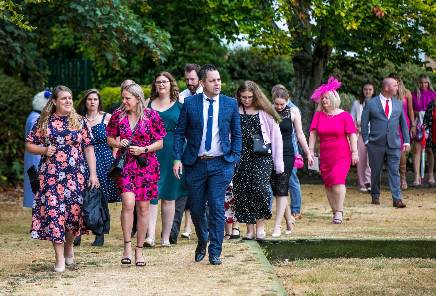 Wedding party walking through a sunny garden path in colorful attire.