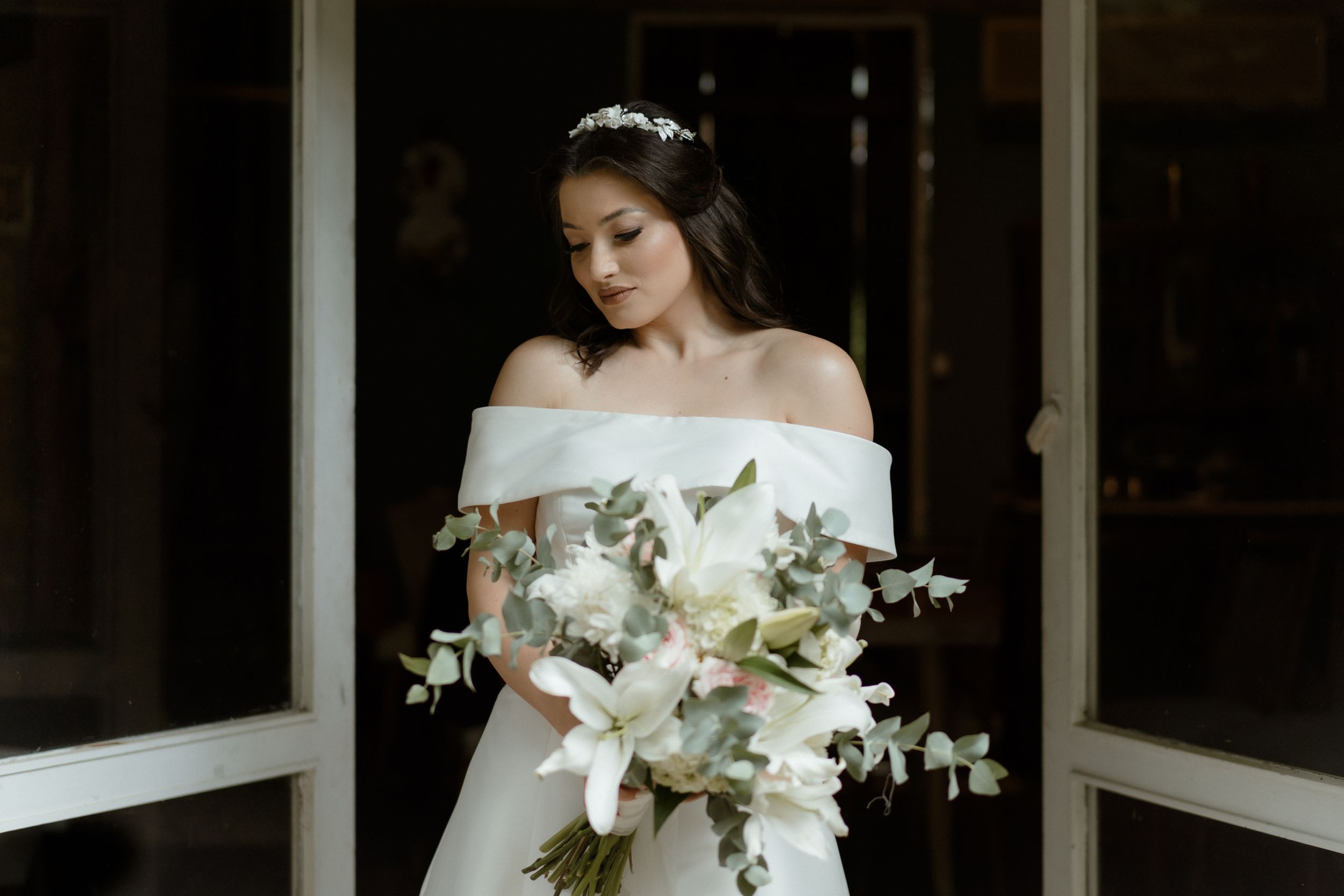 Bride in off-shoulder dress holds a white floral bouquet against a dark background.