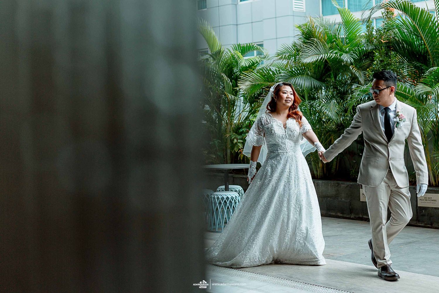 Couple in wedding attire walking hand in hand through modern indoor space with palm plants.