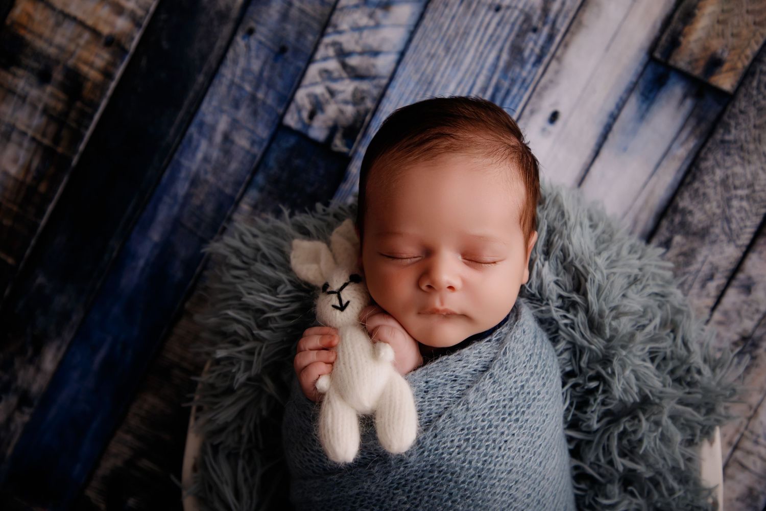 Sleeping newborn wrapped in gray blanket holding small stuffed animal against rustic backdrop.