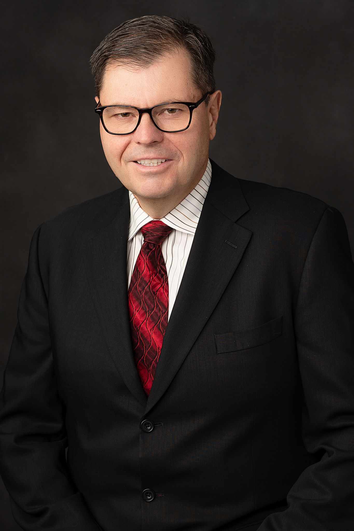 Professional headshot of person in black suit and red tie against dark backdrop.