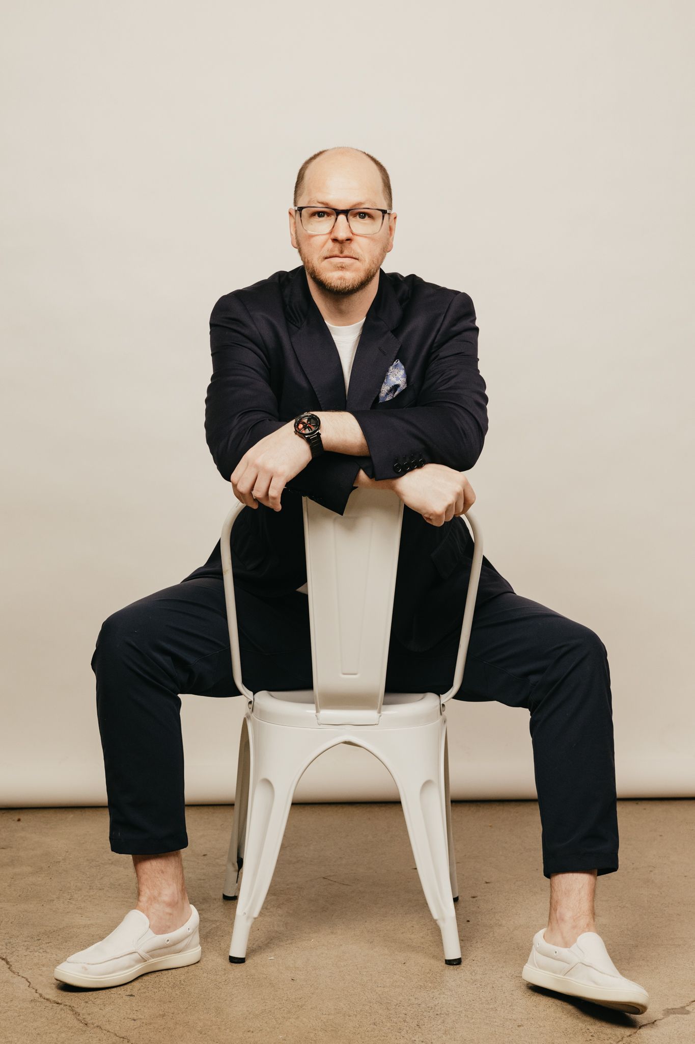Person in black suit sits casually on white metal chair against neutral background.