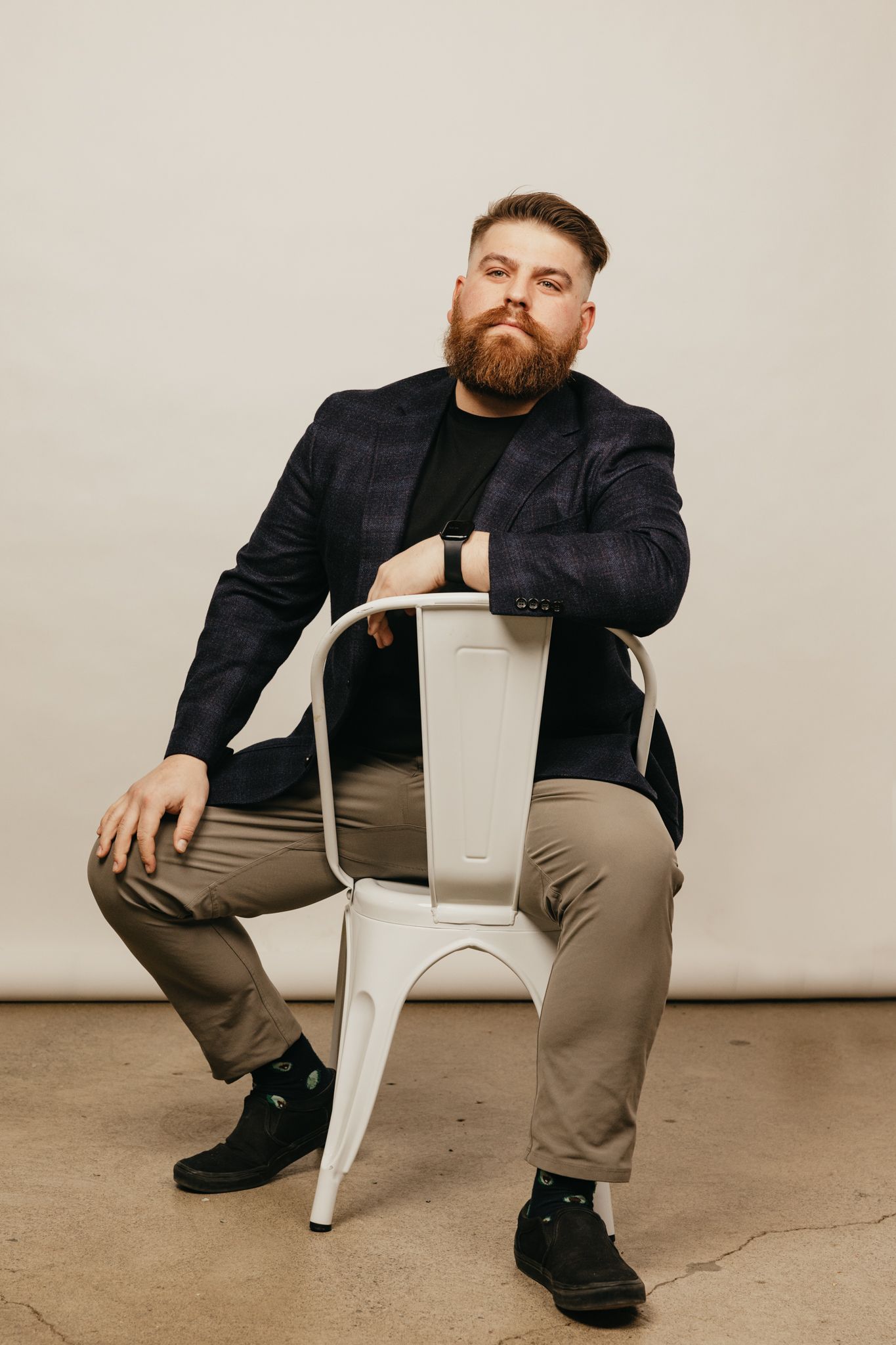 Person in dark blazer and khaki pants poses with white metal chair on concrete floor.