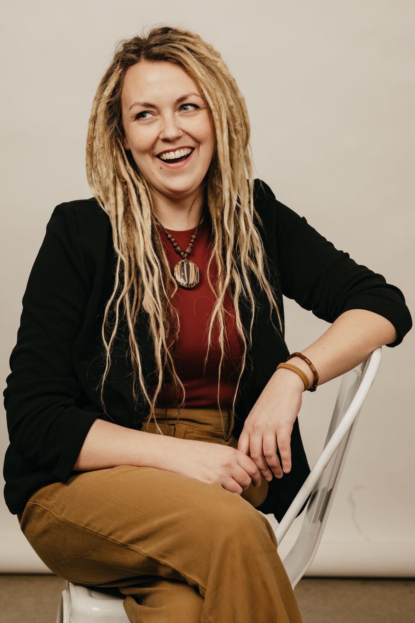 Person in black jacket and brown pants sits casually on white chair against beige backdrop.