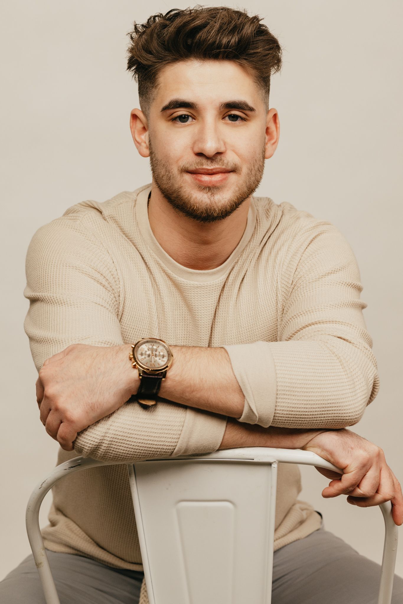 Studio portrait of a person in a beige sweater sitting on a white chair.