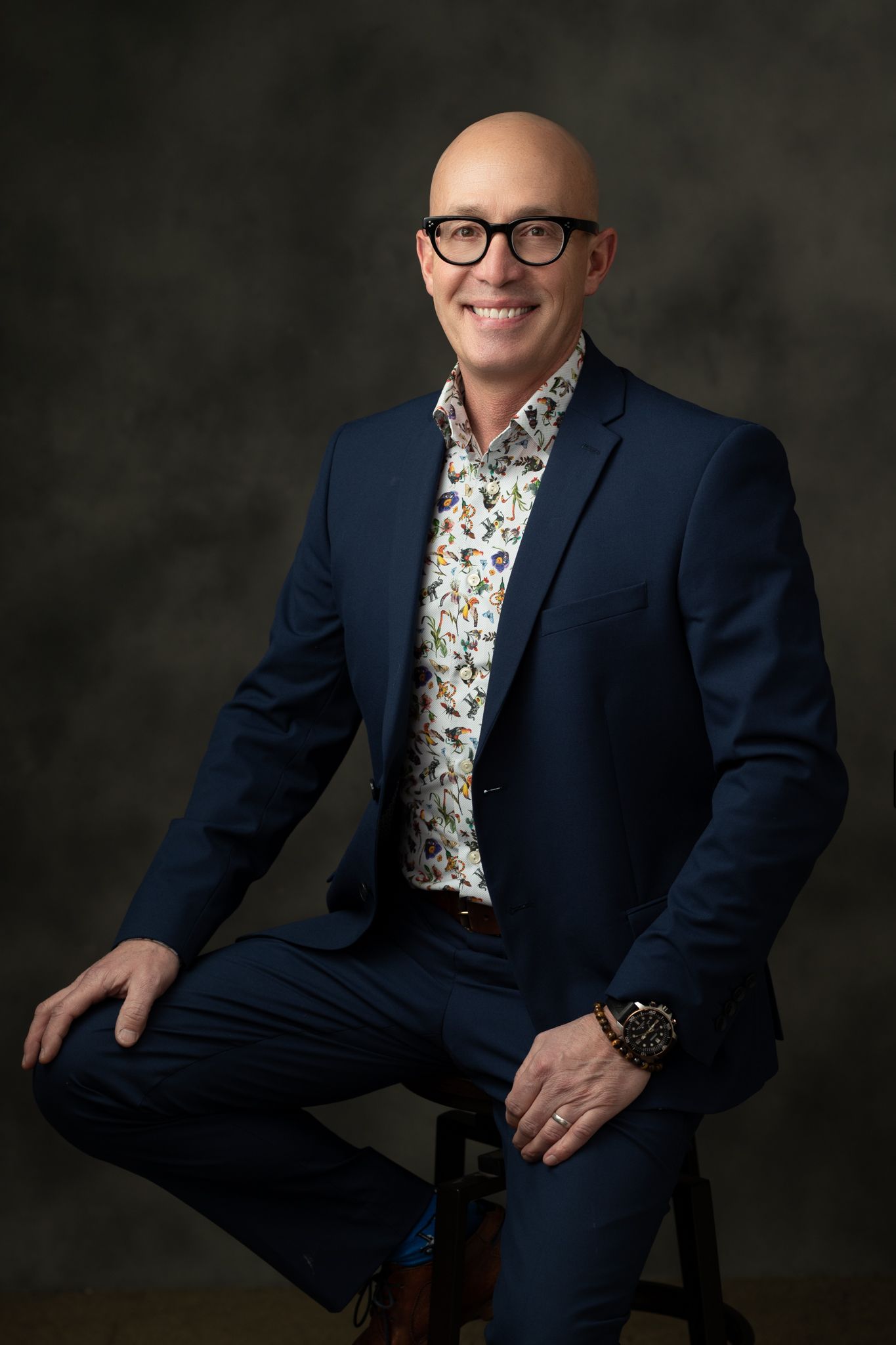 Professional portrait of person in navy suit and floral shirt against dark background.