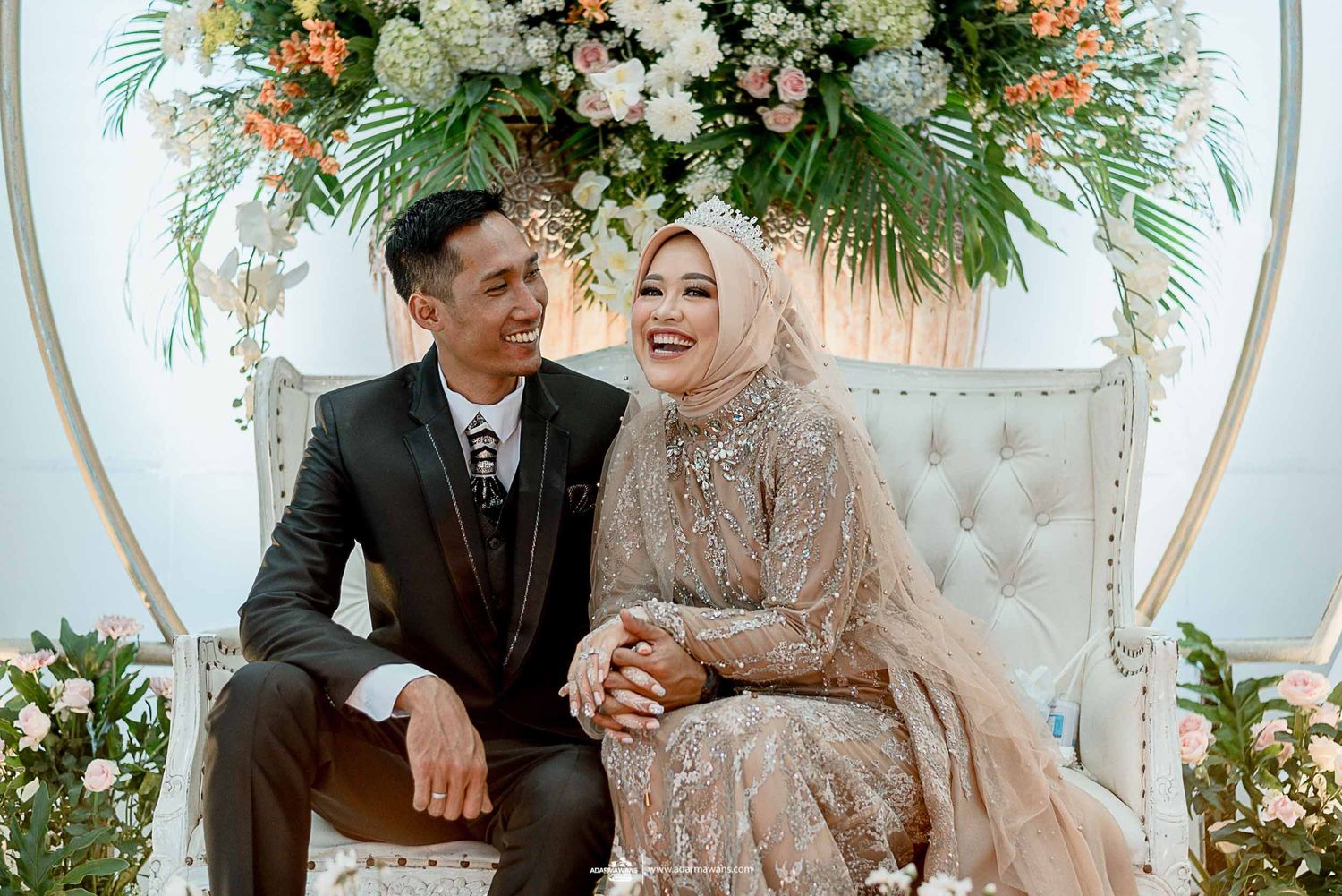 Wedding couple seated on elegant furniture surrounded by floral decorations.