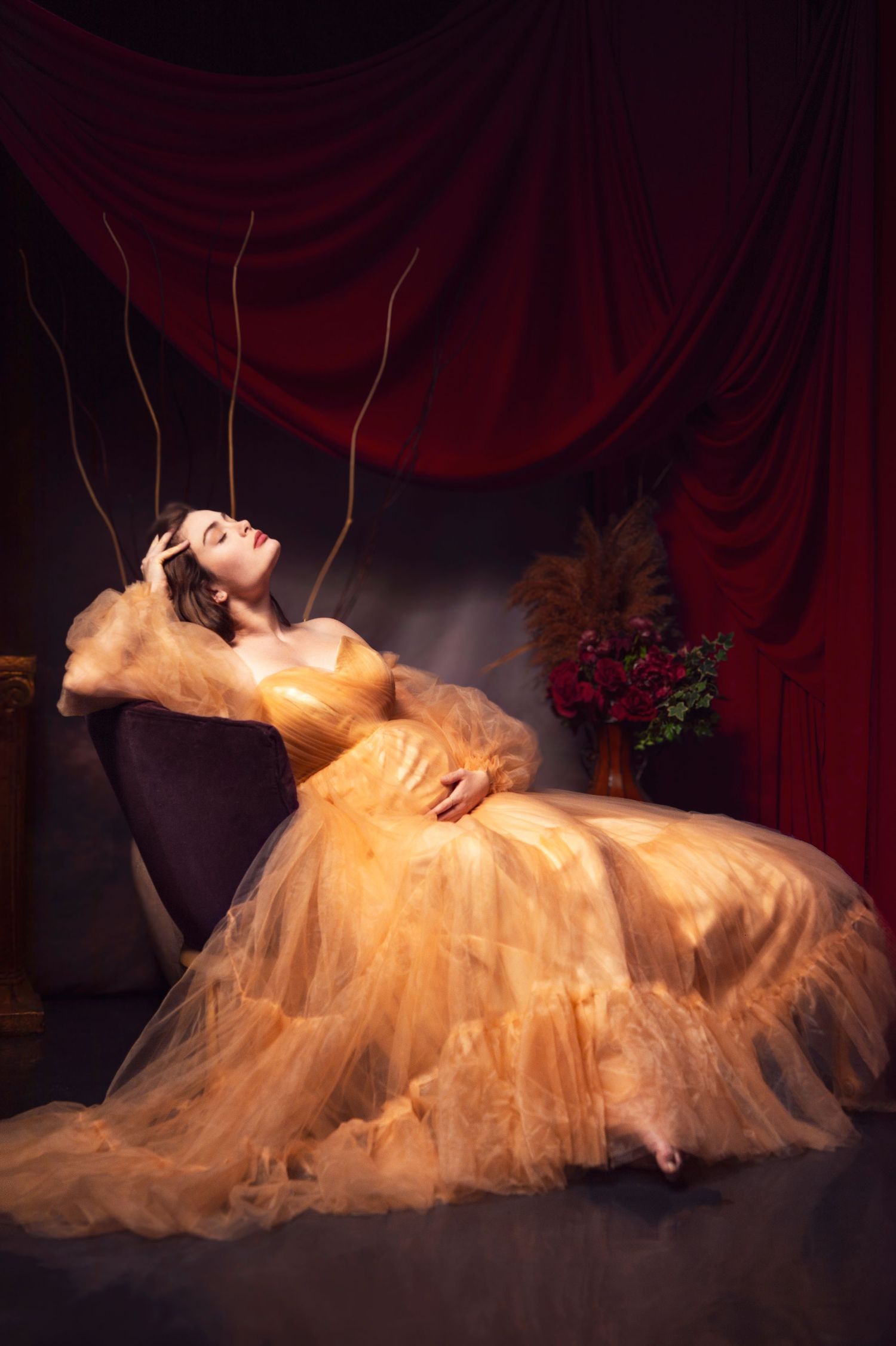 Dramatic portrait of person in golden gown reclining against burgundy curtain backdrop.