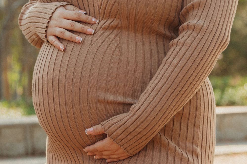 Close-up of pregnant belly in ribbed beige sweater dress with hands cradling bump.