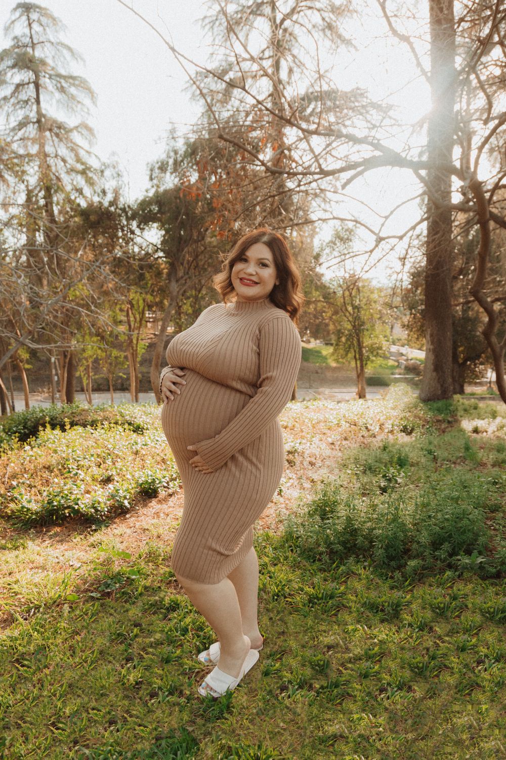 A maternity photoshoot in a wooded park featuring a brown knit sweater dress and white shoes on a sunny autumn day.