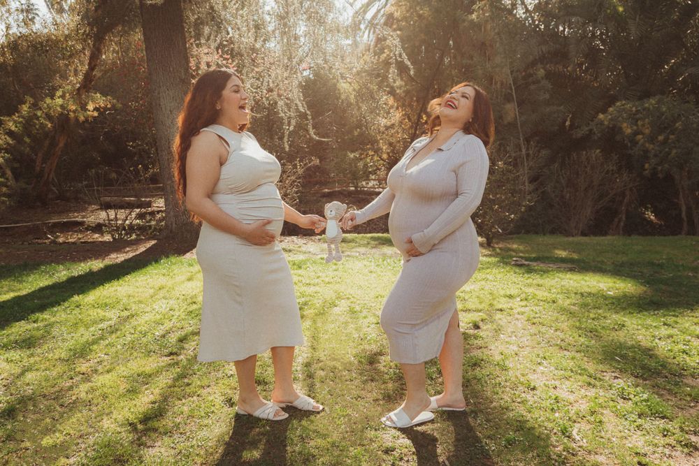 Two pregnant women in white dresses share a laugh in sunlit garden setting.