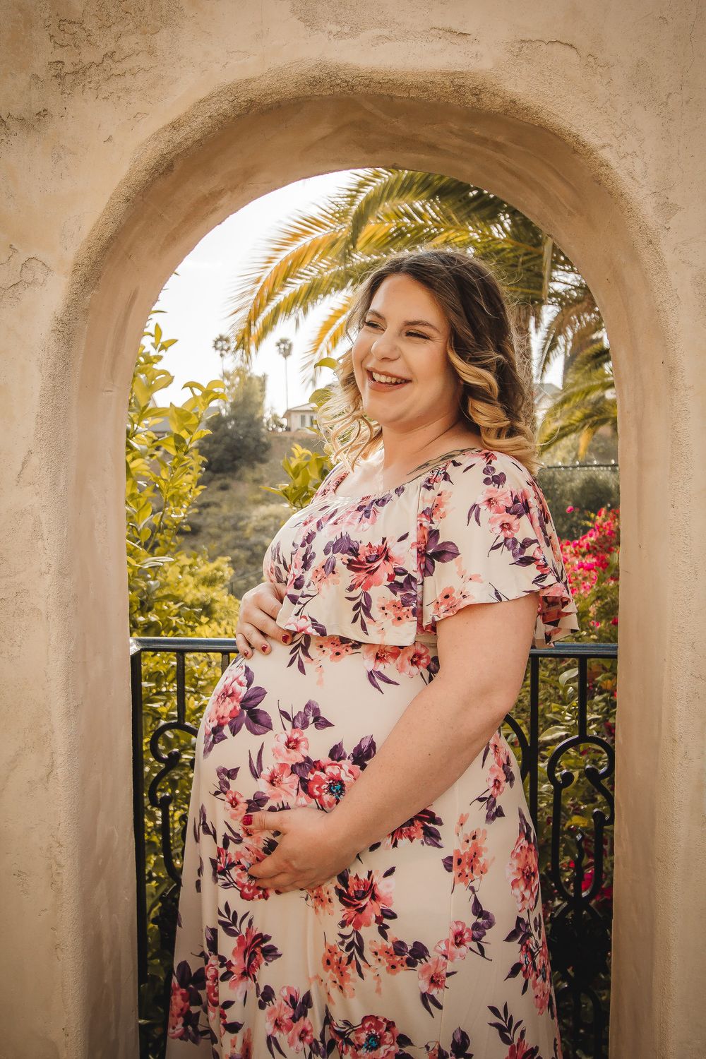 A joyful maternity portrait taken through an arched doorway with palm trees in the background.
