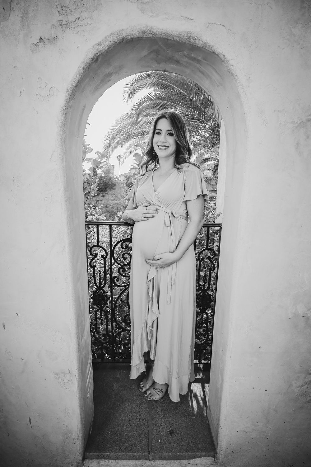 Black and white maternity portrait through an arched doorway with wrought iron details.